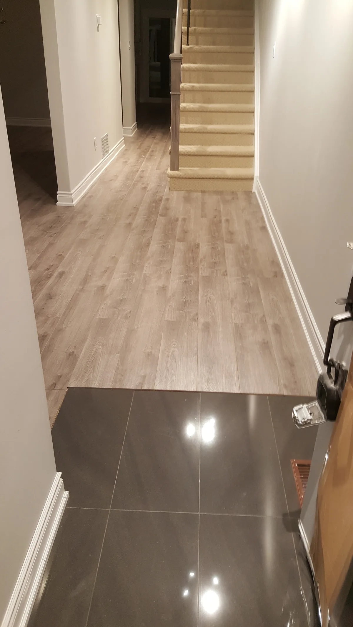 Finished lower-level hallway where vinyl plank flooring meets large-format polished tile, with white baseboards and a carpeted staircase